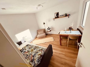 Une pièce de taille modeste comprenant un lit, une petite table et une chaise, un canapé et une étagère ornée d’objets décoratifs.