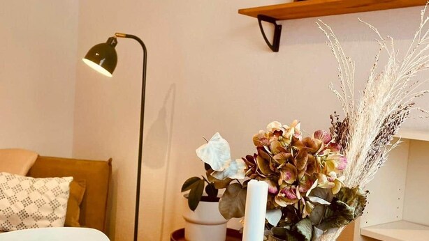 Una sala de estar acogedora con una mesa de centro de madera, un florero de cristal transparente con flores, una lámpara de pie y una estantería con adornos.