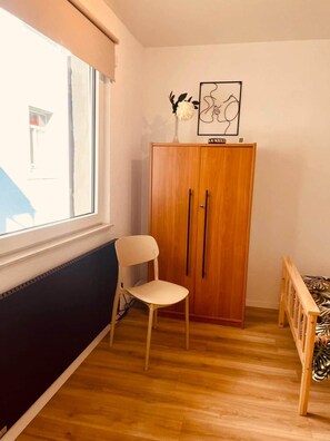 Une pièce comprenant une armoire en bois, une chaise, un lit recouvert d’une couvre-lit à motifs, une fenêtre donnant sur un immeuble et une œuvre d’art encadrée accrochée au mur.