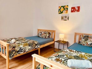 Ein Zimmer mit zwei Holzbettgestellen, jeweils mit blauer Matratze und einem zudeckenartigen Bezug mit tropischen Blättern. Zwischen den Betten steht ein Nachttisch mit einer Lampe.