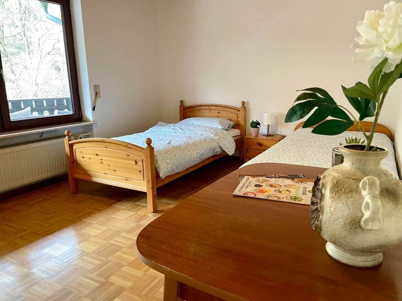 A small bedroom with a wooden bed, a bedside table, a vase with a flower, and a window with a view of trees.