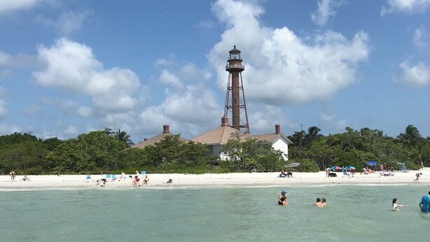Ein Strand mit einem Leuchtturm im Hintergrund, klarem türkisblauem Wasser und Menschen, die schwimmen und in der Sonne entspannen.