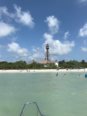 En strand med en fyr i bakgrunden, kristallklart turkost vatten och människor som badar och kopplar av på sanden.