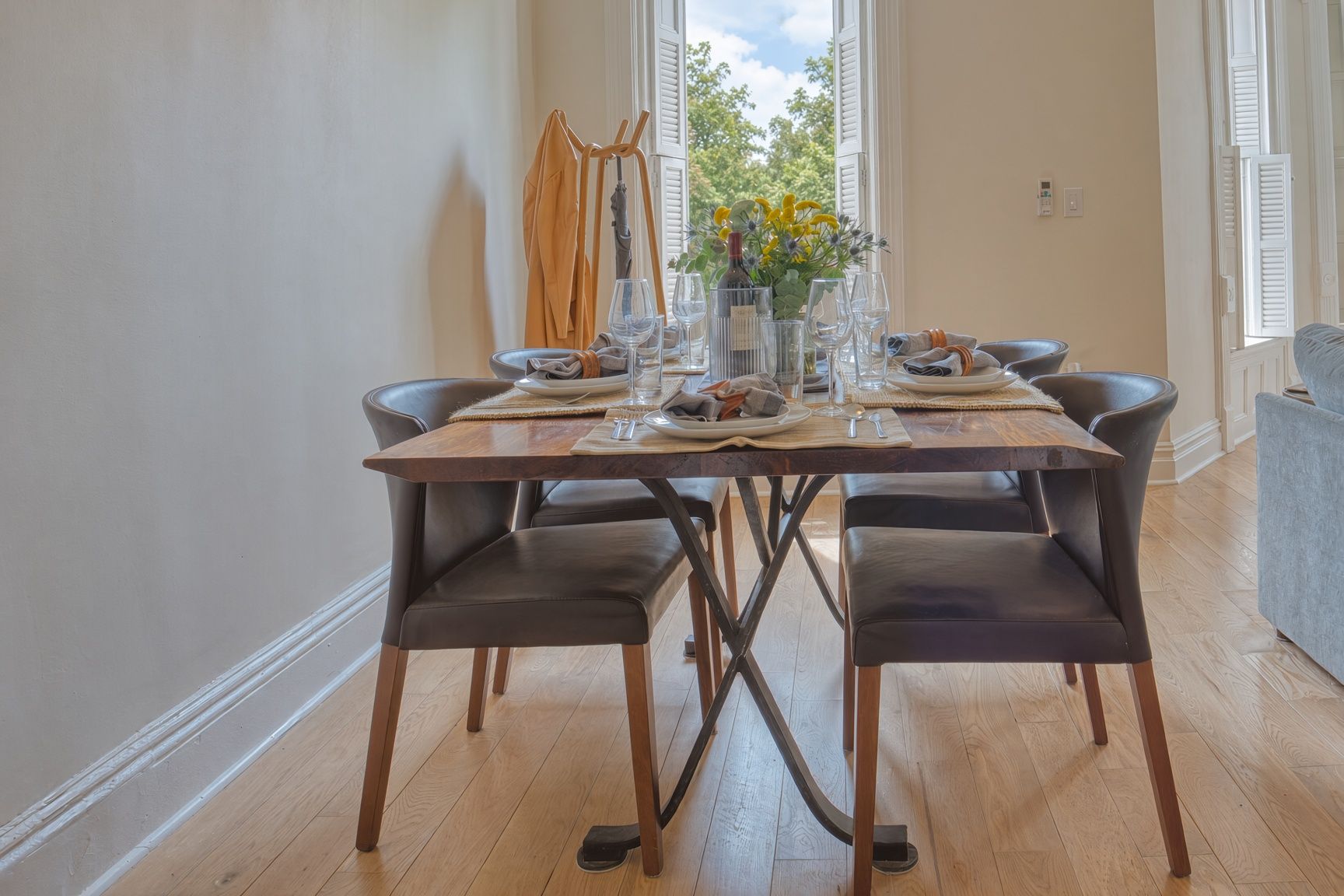 Une table à manger, dressée avec des assiettes, des verres et une bouteille, entourée de chaises, dans une pièce au plancher en bois et offrant une vue par une fenêtre.