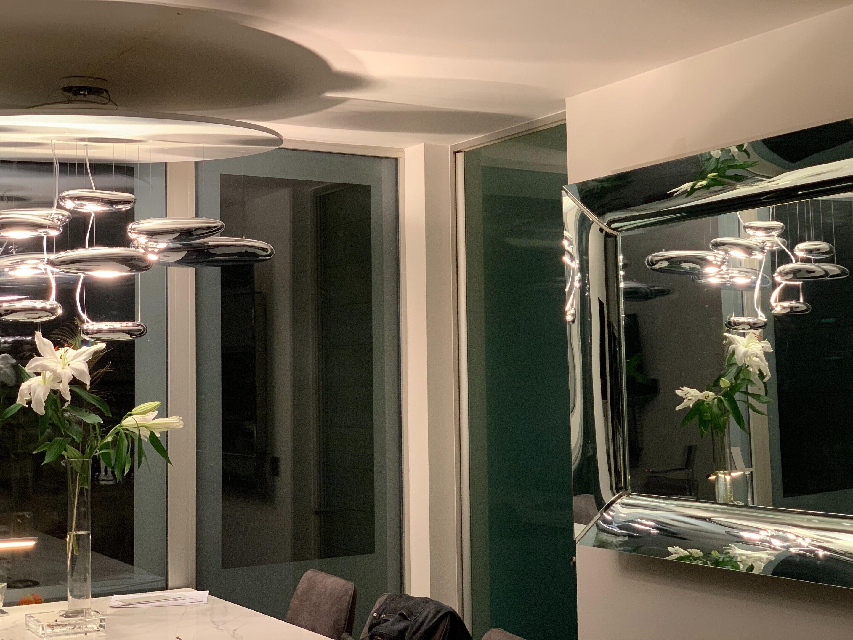 A modern dining area with a long table, chairs, a vase with flowers, and a large mirror on the wall.