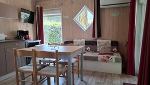 A compact room with a wooden dining table and chairs, a sofa with floral cushions, a television mounted on the wall, and a window with curtains.