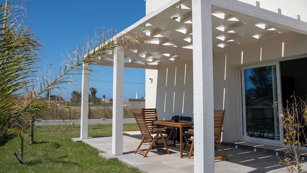 En overdækket udendørs terrasse med et bord og stole, en pergola og havudsigt.