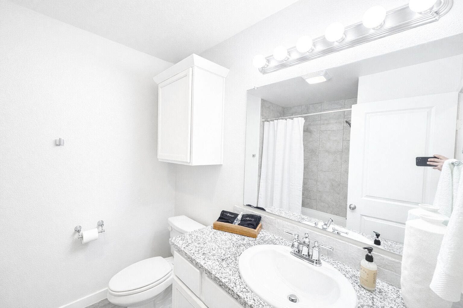 Une salle de bain moderne avec des toilettes blanches, un lavabo surmonté d’un plan de travail en granit, un grand miroir avec éclairage intégré et un espace douche.