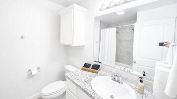 Une salle de bain moderne avec des toilettes blanches, un lavabo surmonté d’un plan de travail en granit, un grand miroir avec éclairage intégré et un espace douche.