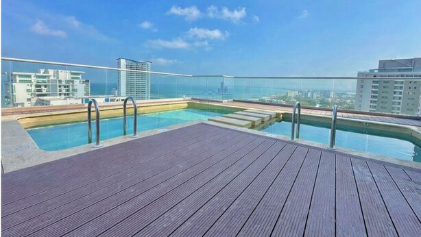 Une piscine sur le toit, dotée d’une terrasse en bois et de balustrades en verre, offrant une vue imprenable sur le paysage urbain et la mer.