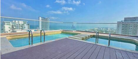 A rooftop pool with a wooden deck and glass balustrades, overlooking a cityscape and the sea.