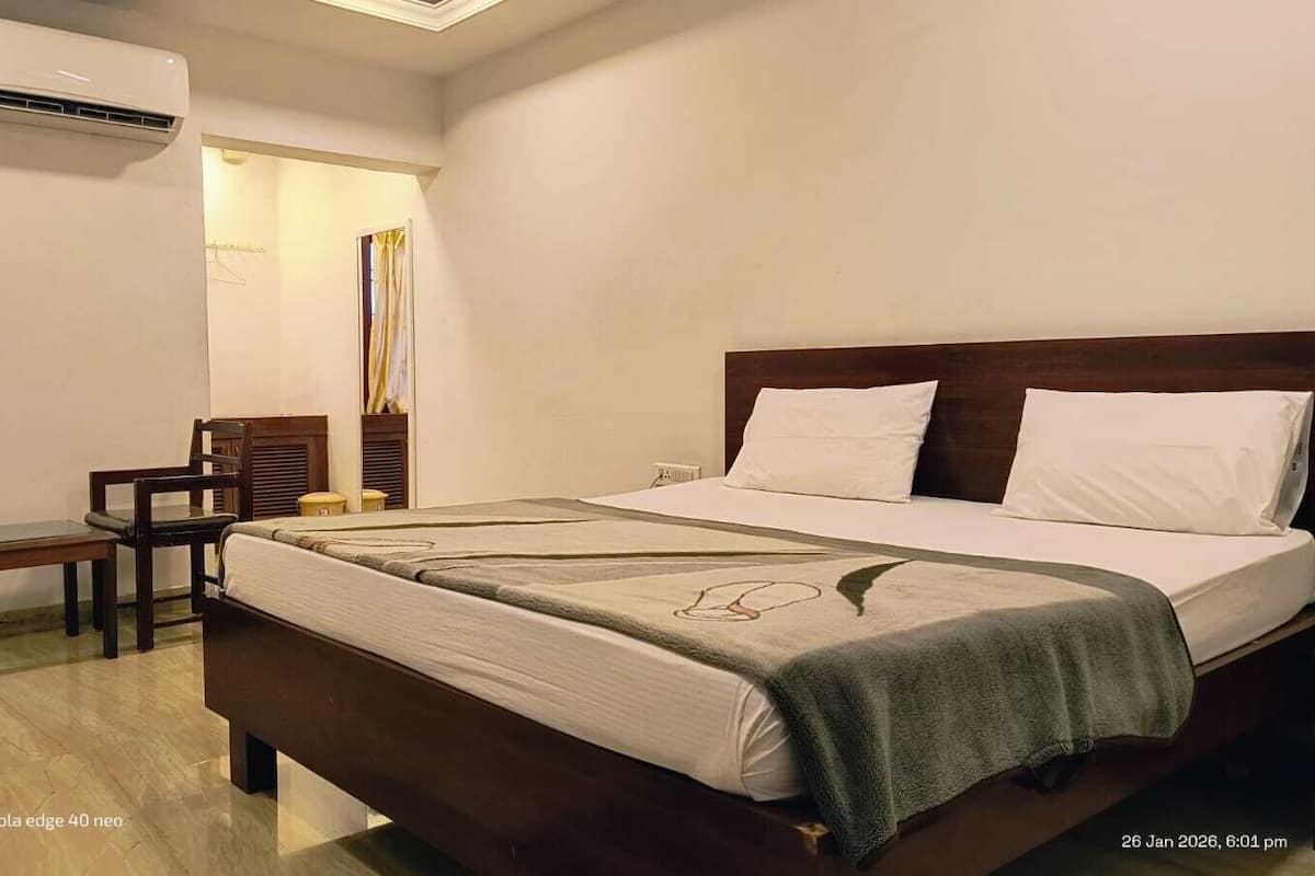Hotelkamer met een bed, een bureau en een stoel. Er is een raam met gordijnen, een wandairconditioner en een houten hoofdeinde.