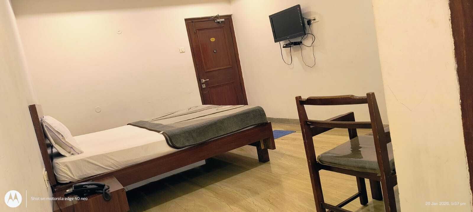 A small, simple room with a single bed, a wooden chair, a television mounted on the wall, and a door.
