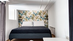 Ein kleines, modernes Schlafzimmer mit einem Bett, einem weißen Sessel, einem kleinen Tisch und einer Wand mit blattmusterigem Tapeten.