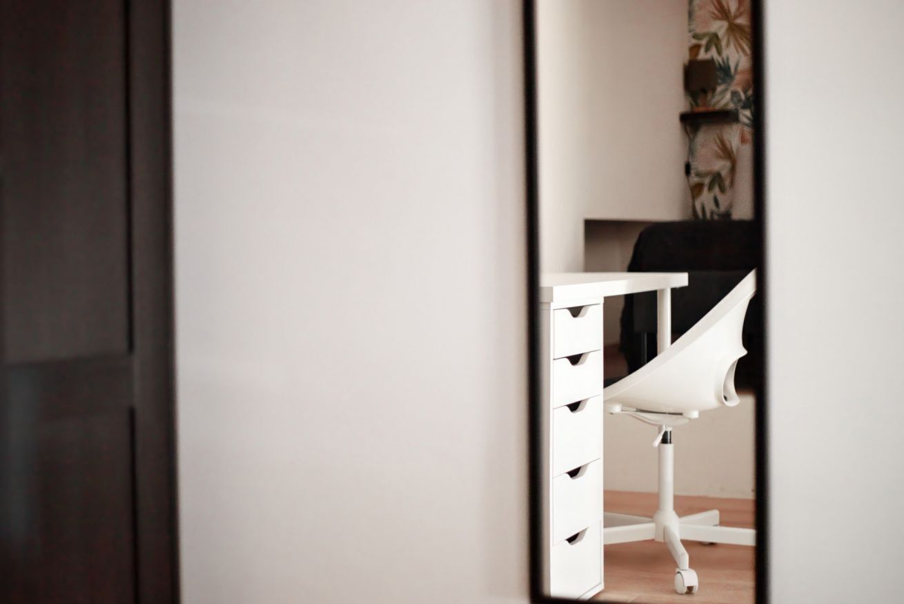 A white desk with drawers and a chair, a dark door, and a wall with a floral pattern.