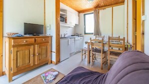 Una sala de estar acogedora con techo de madera, un televisor sobre un mueble de madera, una mesa de comedor con sillas y una zona de cocina con armarios blancos y una ventana.