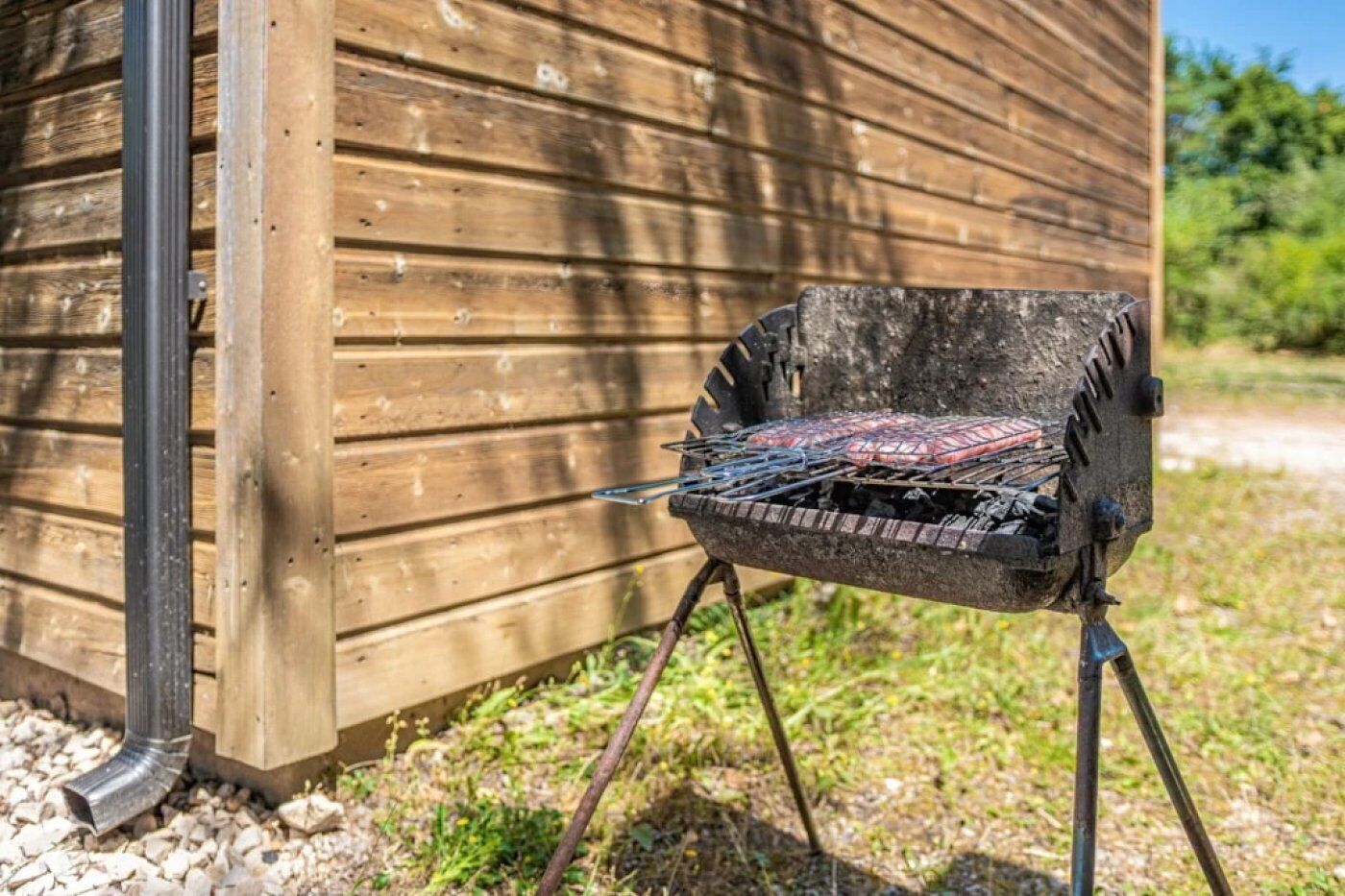 Un barbecue portable doté d’une grille en métal et fonctionnant au charbon de bois, placé sur un support à côté d’un bâtiment en bois.