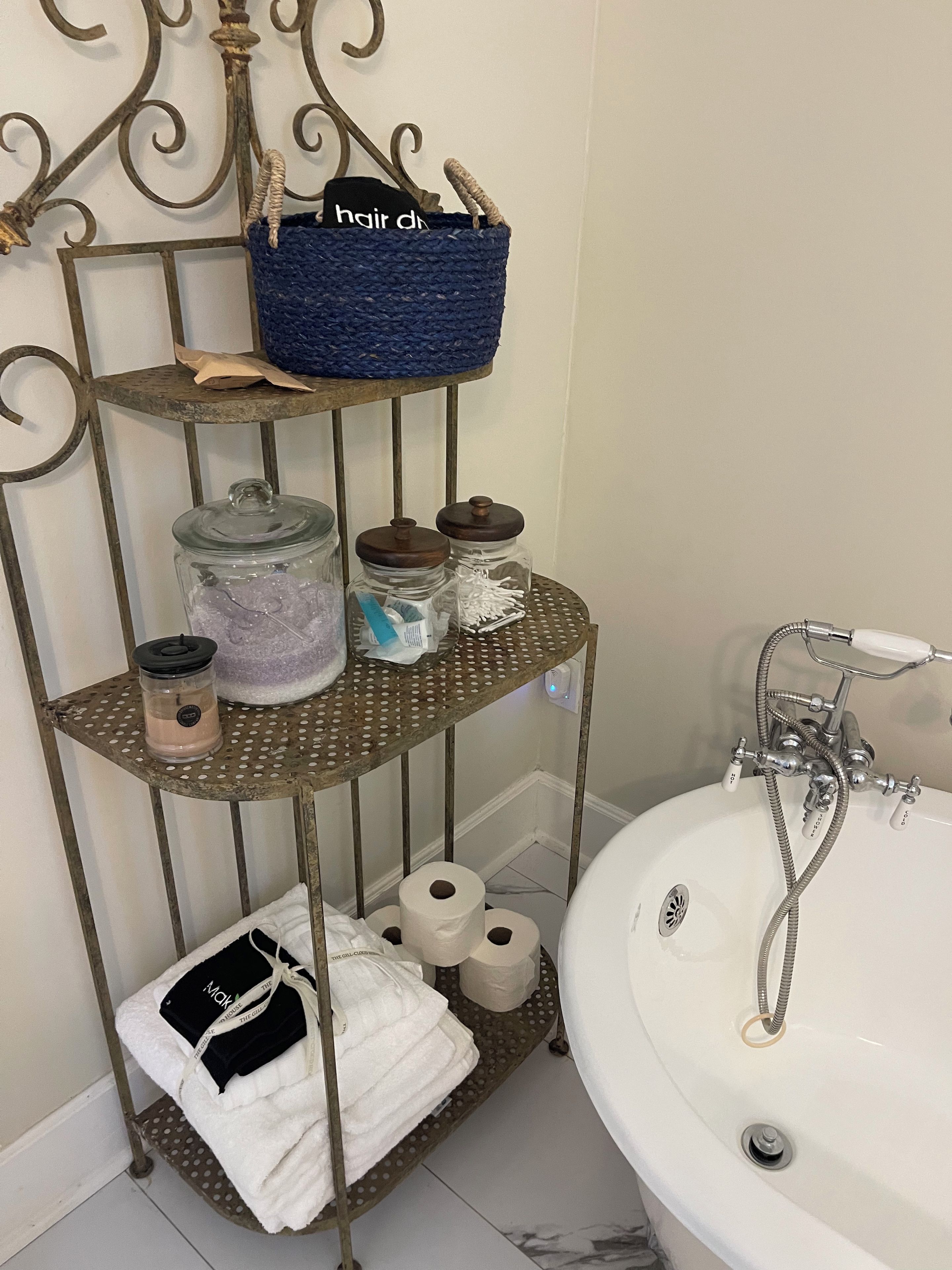 Un tocador de baño con un estante metálico que contiene artículos de aseo, una cesta azul y un lavabo blanco con grifo.