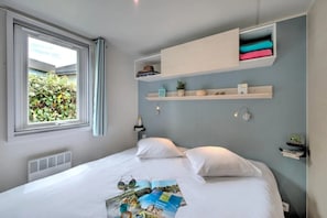 Ein Schlafzimmer mit einem Bett, einem Fenster mit Blick auf Grünflächen, eingebauten Regalen mit Gegenständen und einer Wandleuchte.