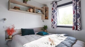 A bedroom with a bed, a window with patterned curtains, a shelf with plants and books, and a vase with flowers. - ZEcamping 4* piscine couverte club enfant jacuzzi locatif TV LV clim (Aureilhan)