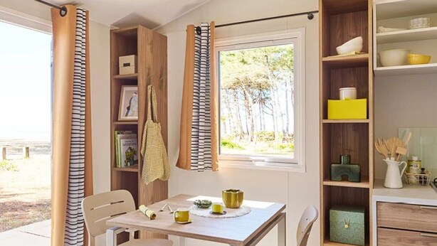 A dining area with a table, chairs, and a window with curtains. There are shelves with various items and a view of trees outside. - ZEcamping 4 étoiles mobil-home TV 16m2 (Morsalines)