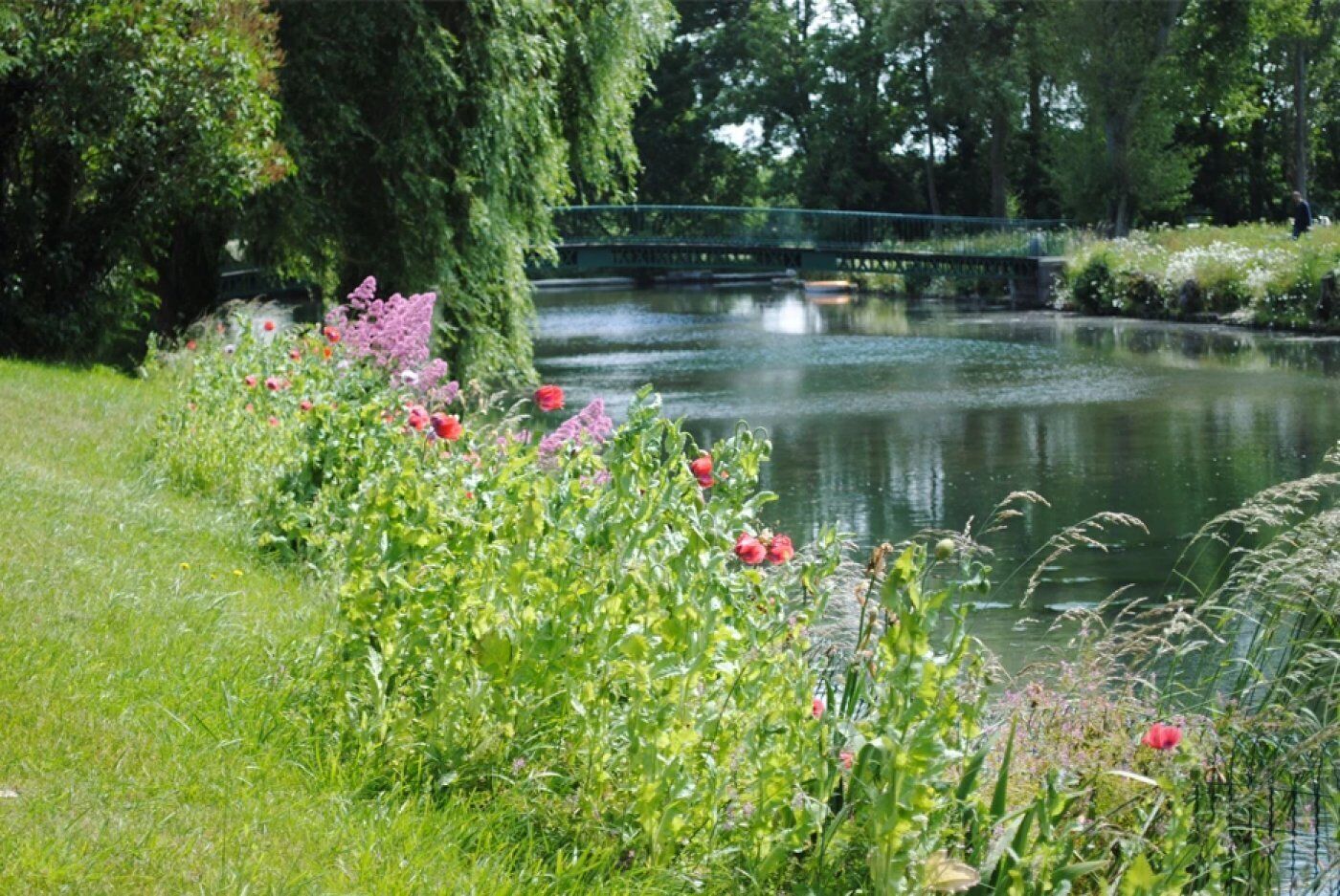 Un jardin paisible au bord de la rivière, agrémenté d’un pont, d’une végétation luxuriante et de plantes en fleurs.