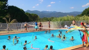 Ein Schwimmbecken mit darin badenden Personen, umgeben von einer Ziegelmauer und Bäumen, mit Bergen im Hintergrund.