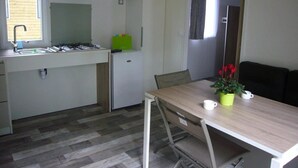 Een kleine keuken met een gasfornuis, spoelbak en een kleine koelkast. Er is een eettafel met stoelen en een potplant.