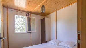 A bedroom with a wooden ceiling, a bed with white bedding, a wardrobe, and a window with a view of trees.