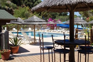 Un área de piscina con sillones de descanso y sombrillas de paja, rodeada de plantas en macetas y una terraza de madera.