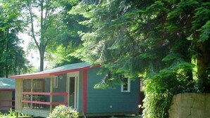 Un piccolo chalet a un piano con finestra a telaio rosso e una porta, immerso nel verde e circondato da un muro in pietra.