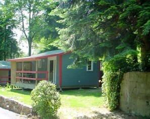 A small, single-story cabin with a red-framed window and a door, surrounded by lush greenery and a stone wall. - ZEcamping 3 étoiles piscine animaux acceptés locatif BBQ clim 30m2 (Mialet)