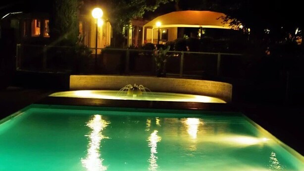 Vista notturna di una piscina con una fontana, circondata da una recinzione e un edificio con finestre illuminate.