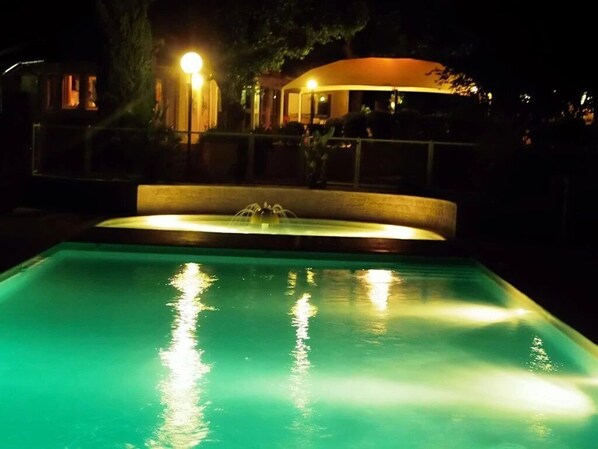 A nighttime view of a swimming pool with a water fountain, surrounded by a fence and a building with illuminated windows. - ZEcamping 3 étoiles piscine animaux acceptés locatif BBQ clim 30m2 (Mialet)