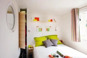 A bedroom with a bed, pillows, a bedside table, a mirror, and a window with curtains. - ZEcamping 3 étoiles piscine chauffée mobil-home 30m2 4 pers (Antonne-et-Trigonant)