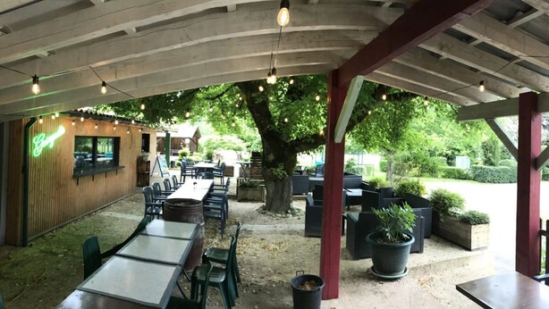 An outdoor dining area with tables and chairs, a wooden structure with a neon sign, and a tree providing shade. - ZEcamping 3* piscine chauffée pataugeoire mobil-home clim 28m2 4 pers (Antonne-et-Trigonant)