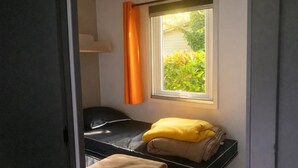 A small, compact room with a bed, a window with curtains, and a view of greenery outside. - ZEcamping 3* piscine chauffée pataugeoire mobil-home clim 28m2 4 pers (Antonne-et-Trigonant)