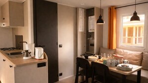 A modern kitchen with a built-in stove, a coffee maker, and a sink. There is a dining area with a table set for two, chairs, and a window with curtains. - ZEcamping 3* piscine chauffée pataugeoire mobil-home clim 28m2 4 pers (Antonne-et-Trigonant)
