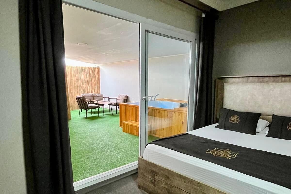 A hotel room with a bed, a wooden headboard, a sliding glass door leading to a balcony with a table and chairs, and a small wooden cabinet.
