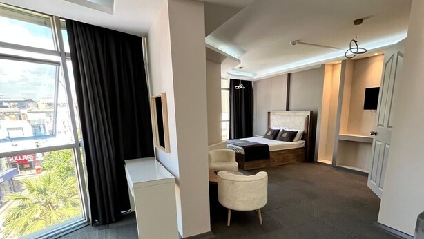 A modern hotel room with a large bed, a seating area with a sofa and armchairs, a small desk, and a large window offering a city view. - ROYAL İNN BORNOVA (İzmir)