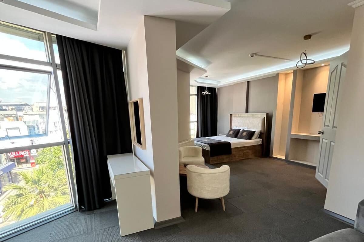 A modern hotel room with a large bed, a seating area with a sofa and armchairs, a small desk, and a large window offering a city view.