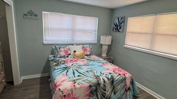 A bedroom with a bed, two windows with blinds, a bedside lamp, and a welcome sign on the wall.