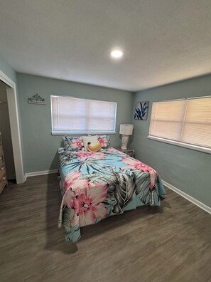 A bedroom with a bed, two windows with blinds, a bedside lamp, and a welcome sign on the wall. - Penguins on the Beach 2BR Beach & Pool  (Cocoa Beach)