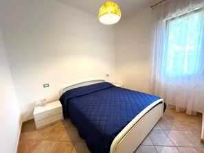 A bedroom with a bed, a nightstand, a lamp, and a window with curtains. - Charming Family Villa With Private Garden (San Michele al Tagliamento)
