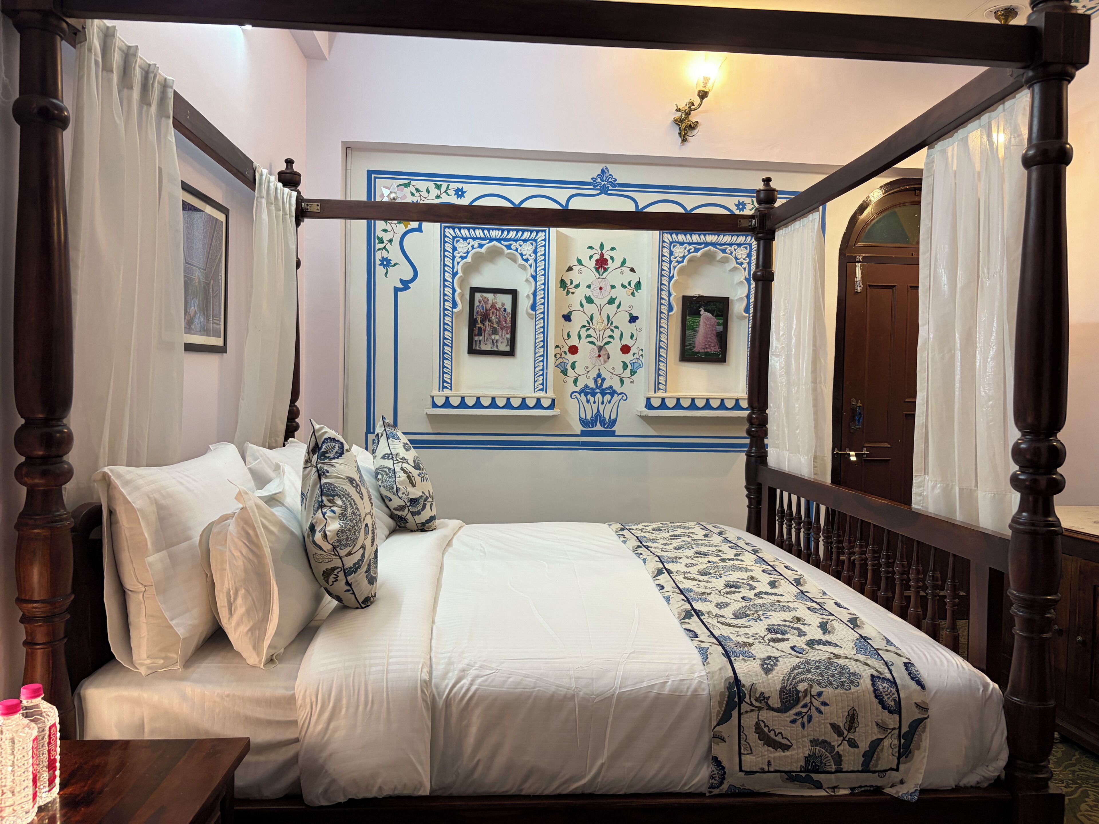 A four-poster bed with a canopy, a bedside table with water bottles, and a wall with decorative blue and white patterns.