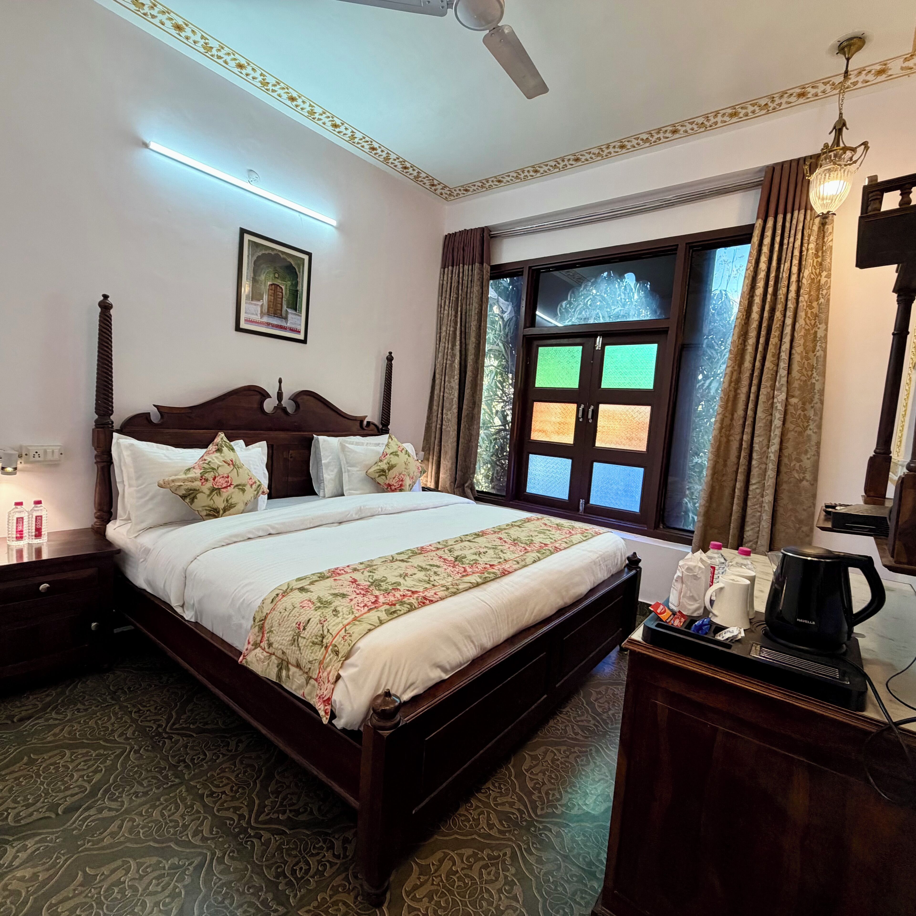A hotel room with a large bed, a ceiling fan, a window with curtains, and a bedside table with a kettle and coffee maker.