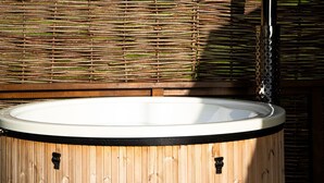 A wooden hot tub with a metal chimney, surrounded by a wooden fence and a tree in the background.