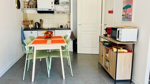 Een kleine keuken met een eettafel, stoelen, een magnetron op een plank, en een deur naar een andere kamer.