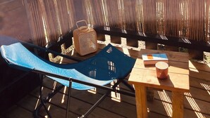 Een houten terras met een blauwe klapstoel, een rieten mand en een kleine tafel met een boek en een mok.