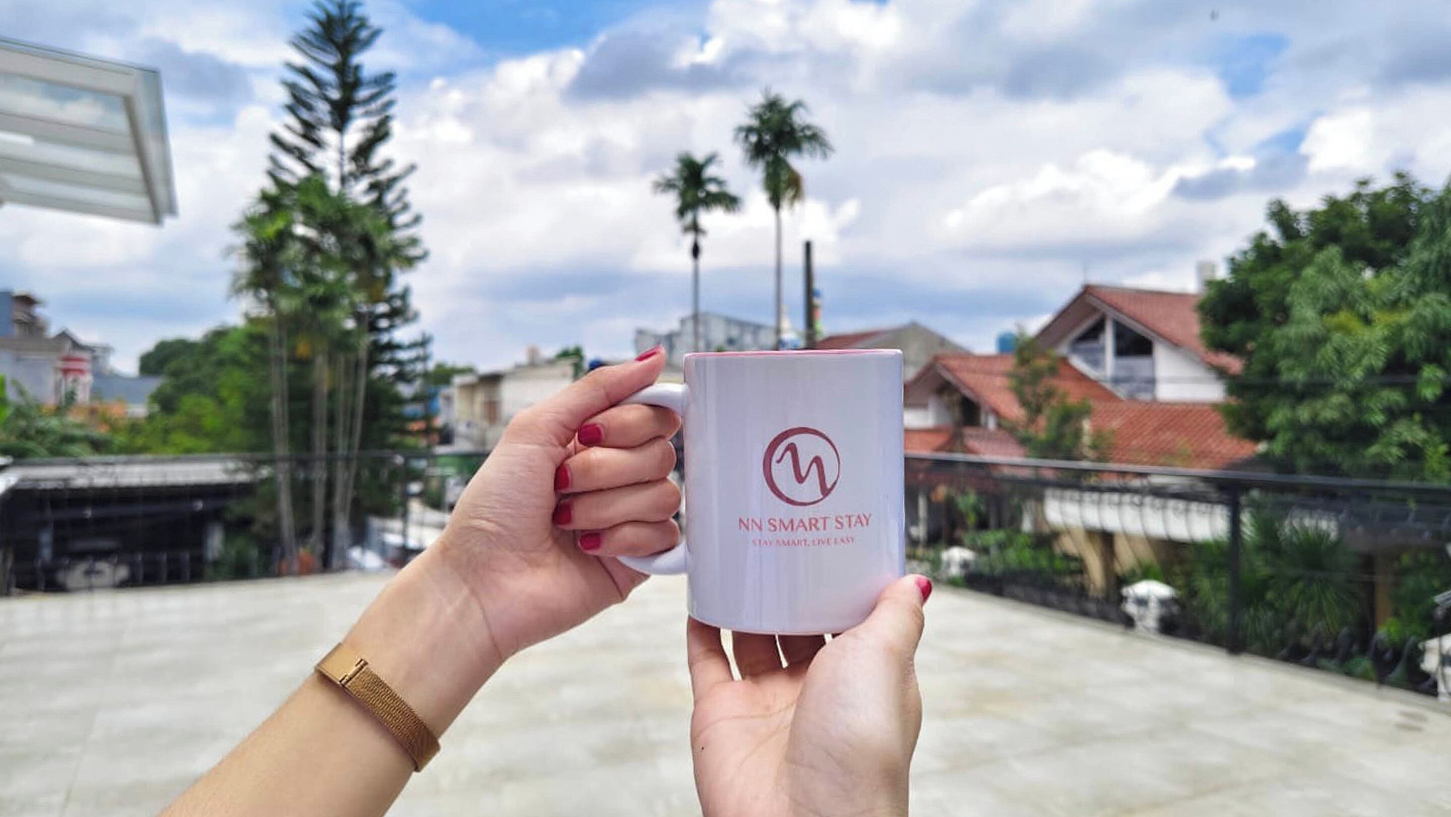 A person holding a mug with the text "NN SMART STAY" on a rooftop with buildings and trees in the background.